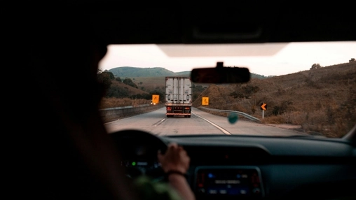 Dashboard view from inside a car trailing a large truck on a two-lane road through dry hillsides at sunset